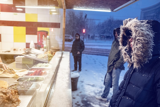 a caucasian girl or woman in a winter jacket with a hood, chooses a street food at a stall in a winter evening city.