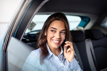 Fototapeta na wymiar Beautiful business woman is using a smart phone and smiling while sitting on back seat in the car. Portrait of beautiful smiling happy woman in back of a car. Beautiful business woman in car.