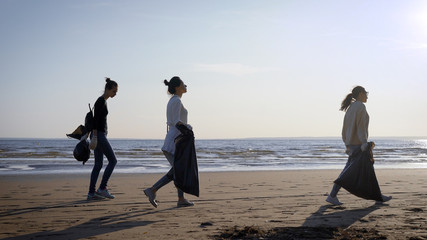female volunteers are walking with plastic bags over wild sand beach in daytime, cleaning city...