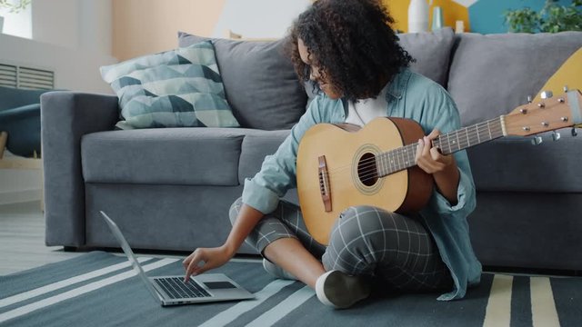 Slow Motion Of Joyful Afro-American Lady Playing The Guitar In House Sitting On Floor Using Laptop Learning How To Play The Musical Instrument Online