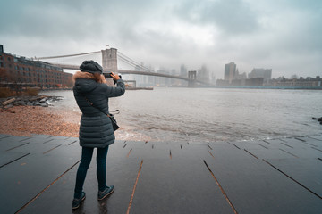 Tourist woman with warm clothes taking a picture of Brooklyn Bridge while sightseeing new york...
