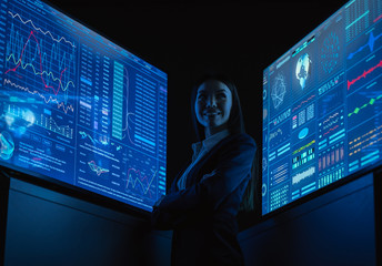 The smiling business lady standing between blue monitors in the dark office