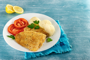 Close-up of a healthy baked battered fish, seasoned with fresh parsley and lemon and accompanied by cooked potatoes and tomato. Blue background. Concept of nutritious food for a healthy diet.
