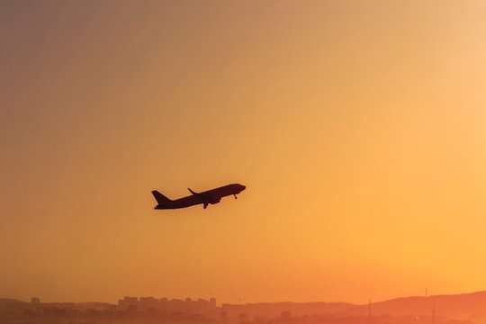 Dark Silhouette Of A Plane Is Gaining Altitude Above A City. Against A Background Of Orange Sky With Smog And Polluted Air.