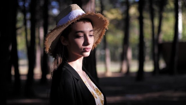wonderful model in yellow straw hat lit by sunlight stands in dense forest against blurry trees slow motion close view