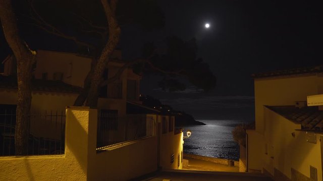 Moonlight reflected in a sea ripple on the shore of a small Mediterranean village. Full moon. Lunar path in the night sea. Yellow light of a lantern on a quiet street leading to the beach. Moon night.