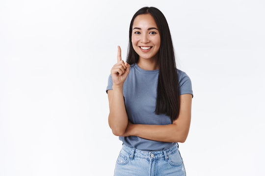 Attractive modern asian brunette woman have good idea, raise finger explain number one rule or showing best choice, indicate up, smiling as telling something important, white background