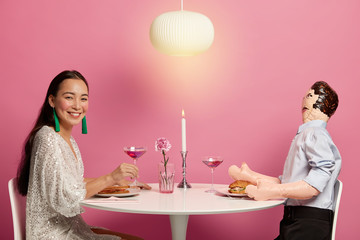 Horizontal shot of woman and inflated doll man have dinner together with candle, drink cocktail, pretend being boyfriend and girlfriend. Asian female dates with unreal layman, pose in restaurant
