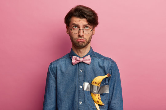 Frustrated unhappy man concentrated above with sad expression, has banana duct tape trend on clothed, wears denim shirt, glasses and bowtie, thinks about something unpleasant, bad mood Modern art.