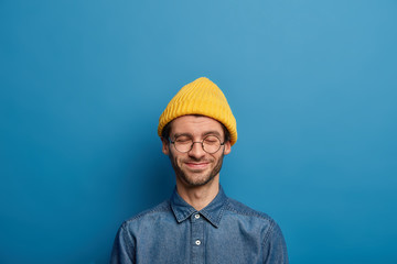 Optimistic Caucaisan man keeps eyes closed, smiles happily, wears yellow headgear and denim shirt, has pleasant thoughts, isolated over blue studio wall. Youth and pleasant emotions concept.