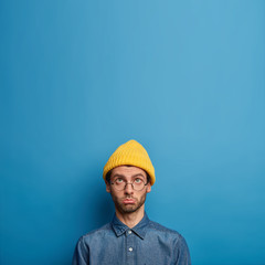 Upset offended man focused above, complains of hard life, wears yellow hat and denim shirt, looks up with sad face, being always pessimistic, isolated over blue wall with copy space for information