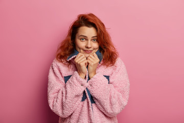Pretty pleased ginger haired girl feels comfort wearing new pink warm coat, looks directly at camera, has dimples on cheeks, poses against rosy background, feels shy in presence of handsome man