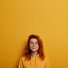 Vertical image of lovely ginger girl looks above, has pleased expression, raises head, enjoys sunny day, wears big glasses and sweatshirt, isolated over yellow background, copy space area for advert