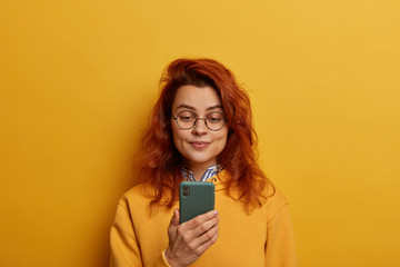 Naklejka premium Ginger young woman holds mobile phone, reads notification, wears round glasses and yellow jumper, connected to wireless internet, isolated over yellow wall. Monochrome. People and technology concept