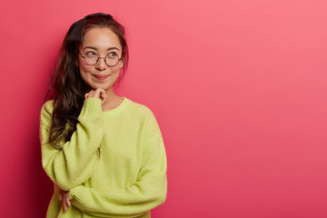 Shot of attractive thoughtful girl recalls first romantic date with boyfriend, keeps hands gently under chin, has dreamy happy expression, wears casual clothes, models over rosy wall, copy space