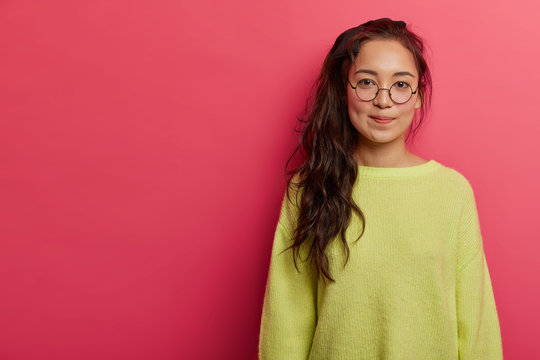 Calm serious woman with long pony tail, wears transparent glasses, green jumper, looks attentively at camera, poses in pink studio, decides on something important, has clever intelligent look