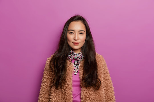 Serious dark haired woman in brown coat, has healthy skin, minimal makeup, looks with charming expression, has straight gaze at camera, isolated over lilac background. Asian female in outerwear
