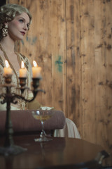Retro 1920s fashion woman sitting on sofa beside table with candlestick and glass of champagne.