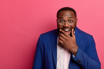 Joyful black man boss holds chin and laughs happily, dressed formally, has pleased funny...