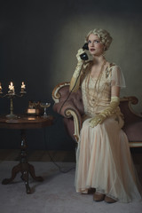 Retro 1920s fashion woman on the phone while sitting on sofa beside table with candlestick and...