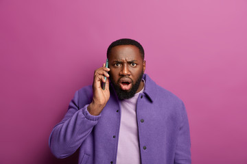 Shocked black man stares in panic, learns bad news during telephone call, gasps from wonder, frowns...