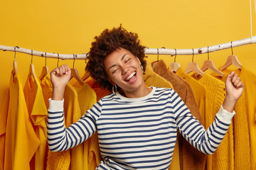 Overjoyed woman shopaholic makes victory dance against clothes rack, happy to buy various clothes,...