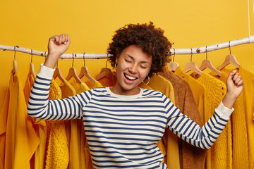 Carefree positive dark skinned woman dances happily, poses near clothes rack, rejoices day off and...