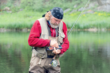 a man with a beard and a bondana, against the backdrop of nature, in fishing equipment with a fishing rod in his hands, selects a bait from a box with tackle