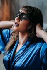 Fototapeta na wymiar Sexy sensual seductive woman in an expensive blue revealing robe and masquerade mask in a dark bedroom with textured walls, self-love, lust, strong independent girl.