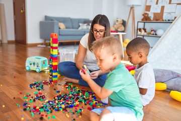 Obraz premium Young beautiful teacher and toddlers playing with building blocks around lots of toys at kindergarten