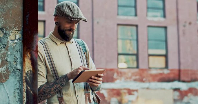 Caucasian young stylish hipster with tattoos and beard standing outdoors and using tablet device, tapping and scrolling.