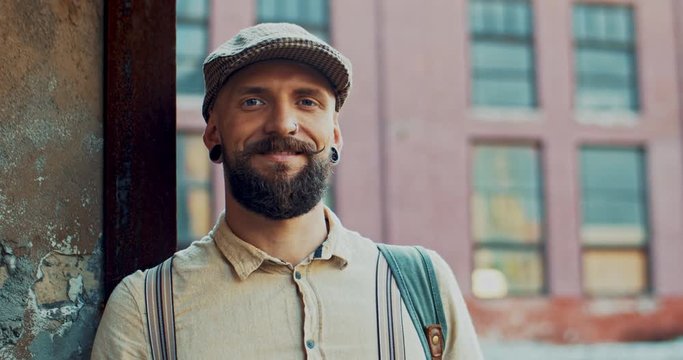 Close up of the young handsome Caucasian hipster in cap and with beard looking at the side, then turning face to the camera and smiling. Portrait.