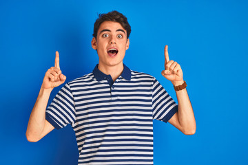 Teenager boy wearing casual t-shirt standing over blue isolated background smiling amazed and surprised and pointing up with fingers and raised arms.