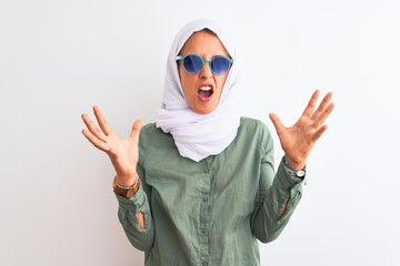 Young Arab woman wearing hijab and summer sunglasses over isolated background crazy and mad shouting and yelling with aggressive expression and arms raised. Frustration concept.