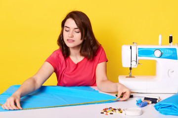 Close up portrait of female dressmaker using sewing machine, sewer sewing dress or shirt from blue fabric, fashion designer taking measurments, looks concentrated, isolated over yellow background.