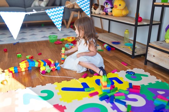 Adorable blonde toddler playing with building blocks around lots of toys at kindergarten