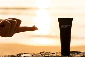 The silhouette of a young woman's hand with a lotion on the finger and a body lotion bottle packaging on the beach in the morning with the light of the sunrise in the background. For Advertisement.