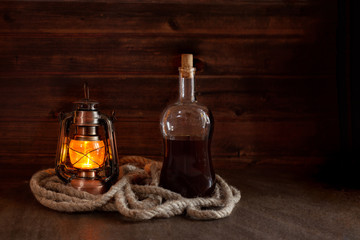 bottle with strong alcohol, rum, whiskey next to an old burnt lantern on a wooden background - obrazy, fototapety, plakaty
