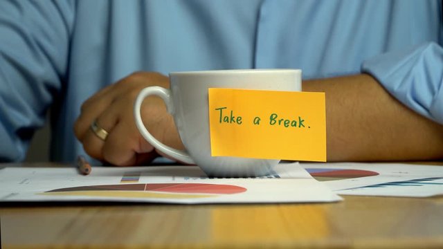 Hand man holding a coffee cup and put it down on the Annual report on coffee cup have a sticky note that says take a break. The concept of encouragement for colleagues