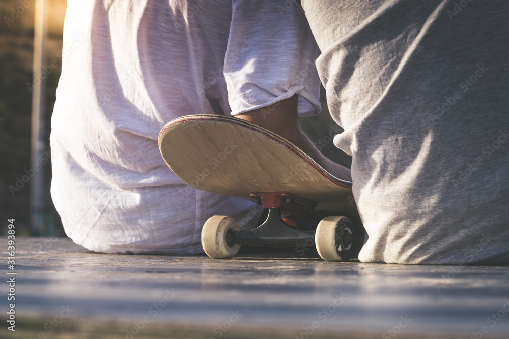 Wall mural Close up back view of two teens sitting on half pipe ramp with skateboard between them. Friendship at the skatepark.Youth togetherness, sport, joy, positive and carefree concept.