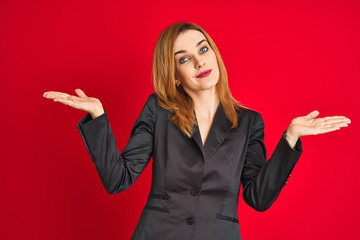 Young caucasian business woman wearing a suit over isolated red background clueless and confused expression with arms and hands raised. Doubt concept.