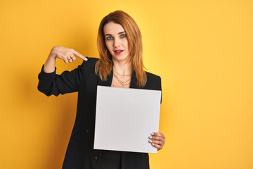 Young redhead businesswoman with clear eyes holding banner over isolated yellow background with surprise face pointing finger to himself