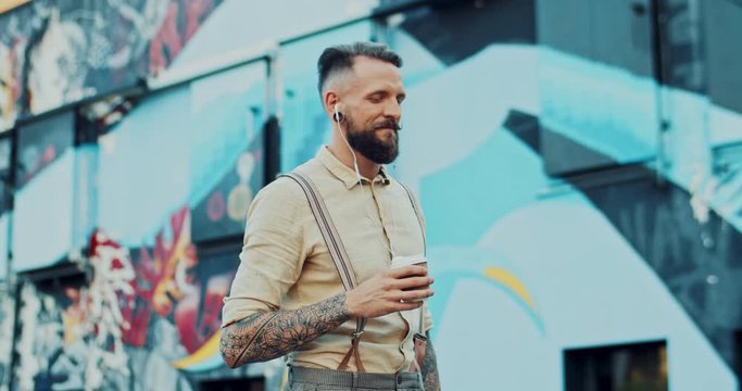 Young Caucasian man with tattoos walking the street at the graffity wall with a cup of coffee to-go and starting to use smartphone. Outdoor.