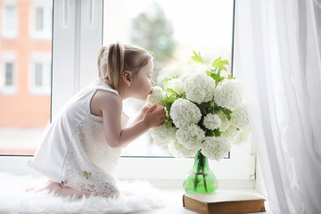 A little girl is sitting on the windowsill. A bouquet of flowers in a vase by the window and a girl...