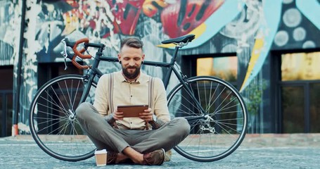 Stylish Caucasian young guy hipster sitting on the ground of the payment at his bicycle and graffity wall and tapping or scrolling on the tablet computer.