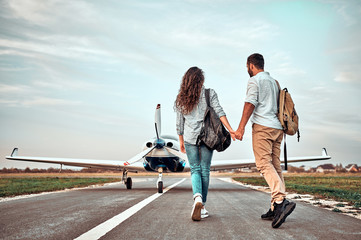 Naklejka na ściany i meble Back view of beautiful romantic couple holding hands while walking on take-off ground near the aircraft
