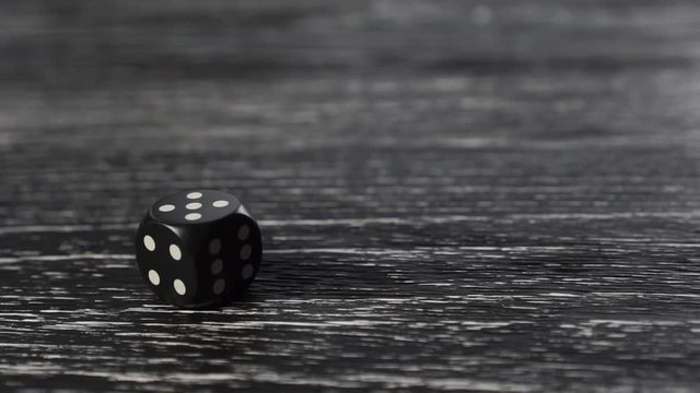 The black dice rolls, spins and stops with the number 5 on top. Dark textured old wooden table. The idea of a good move and good luck in the game and business. Slow motion.