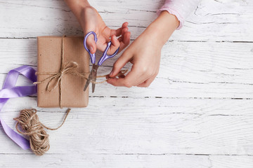 Obraz na płótnie Canvas Child is wrapping gift box in brown kraft paper. Present on white wooden background. Kid cuts ribbon with lilac scissors. Close up of hands. Flat layout. Happy spring holidays. Copy space.