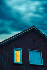 Criminal with pistol in ominous house with illuminated window under stormy sky at dusk.