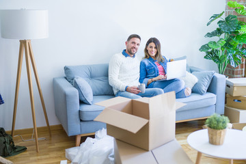 Fototapeta na wymiar Young beautiful couple sitting on the sofa drinking cup of coffee using laptop at new home around cardboard boxes